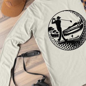 May include: Cream long-sleeved shirt with a black graphic of a golfer swinging a club inside a golf ball design. The design includes a golf course scene with trees and a golf ball. A brown cap and camera are also visible.