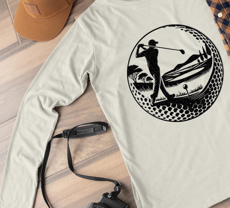 May include: Cream long-sleeved shirt with a black graphic of a golfer swinging a club inside a golf ball design. The design includes a golf course scene with trees and a golf ball. A brown cap and camera are also visible.