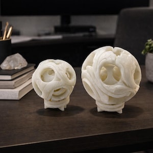 May include: Two white, carved stone spheres with detailed, openwork patterns. They are displayed on a dark wooden desk, with a laptop, a potted plant, and office supplies nearby. The spheres have a distinctive, artistic appearance.
