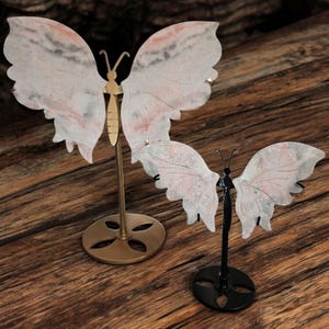 May include: Two decorative butterfly figurines on stands. The larger butterfly is pink and white with a gold stand. The smaller butterfly is white with a black stand.