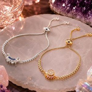 May include: Two adjustable slider bracelets with box chains and beaded sections. One silver-toned bracelet features a crescent moon charm with blue and clear stones. The other gold-toned bracelet features a sun charm with clear stones.