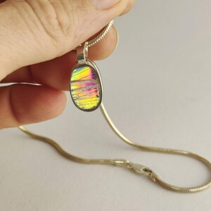 Puede incluir: Collar con colgante de plata con un colgante ovalado iridiscente. El colgante muestra un espectro de colores, incluyendo amarillo, rosa y morado. La cadena es una fina cadena de serpiente plateada con un cierre.