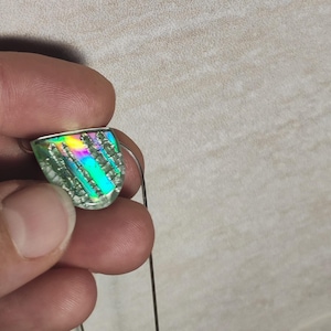 Puede incluir: Un colgante de vidrio dicróico con un marco de metal plateado. El colgante tiene forma de semicírculo con una superficie texturizada y muestra un arcoíris de colores. Se adjunta una fina cadena plateada.