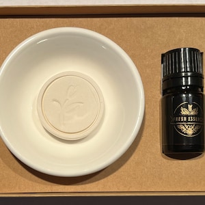 May include: A white ceramic diffuser dish with a white diffuser disc and a small black bottle of essential oil with a gold label that says "Fresh Essences".