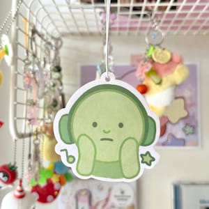 May include: A light green sticker of a cartoon character wearing headphones, with hands on its face, and a sad expression. The sticker is suspended by a white string. Other items are visible in the background.