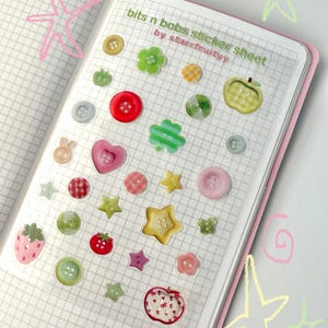 Può includere: Un foglio di adesivi con vari disegni colorati di bottoni e forme, tra cui stelle, fiori e frutta. Il foglio è su carta a quadretti, con il testo "bits n bobs sticker sheet" e "by starrfruityy". Gli adesivi sono in tonalità di rosso, verde, rosa e giallo.