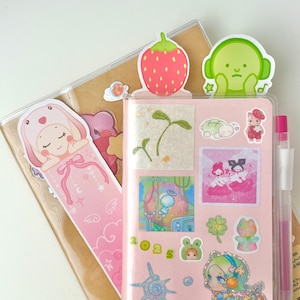 May include: A pink planner with various stickers, including a strawberry, a green character with headphones, and a bunny bookmark. The planner also has a pen and a clear cover.