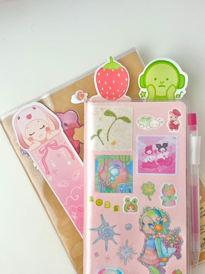 May include: A pink planner with various stickers, including a strawberry, a green character with headphones, and a bunny bookmark. The planner also has a pen and a clear cover.