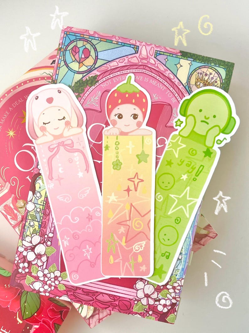 May include: Three colorful bookmarks featuring cute cartoon characters. One is pink with a bunny hat, another is yellow with a strawberry hat, and the last is green with a headphone design. The bookmarks are on top of a book with floral and star designs.