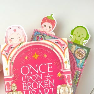 May include: A stack of books with decorative bookmarks. The book covers are pink with the title "ONCE UPON A BROKEN HEART". The bookmarks feature cute cartoon characters, including a bunny, a strawberry, and a green character with headphones.