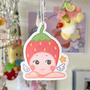May include: A strawberry-shaped air freshener with a cartoon face, wings, and a star. The air freshener is red with a green top, suspended by a white string. Other keyrings and decorations are visible in the background.