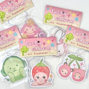 May include: A collection of air fresheners with various cartoon designs, including a monkey, bunny, green character, strawberry fairy, and cherry pair. Each air freshener is packaged with a pink card that reads "Lucky Charm Air Freshener!"