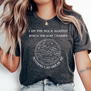 May include: A dark gray t-shirt with a white graphic of a wave crashing against a rock with the text "I am the rock against which the surf crashes. Nothing can break me."