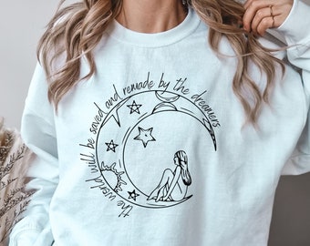 May include: A light blue sweatshirt with a black graphic of a crescent moon with stars and a woman sitting on it. The text "The world will be saved and remade by the dreamers" is written around the moon.