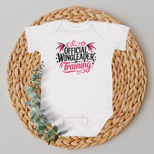 May include: White baby onesie with the text "OFFICIAL WINGLEADER IN TRAINING" in black and pink. The design includes bat wings and hearts. The onesie is displayed on a woven, circular placemat with green foliage.