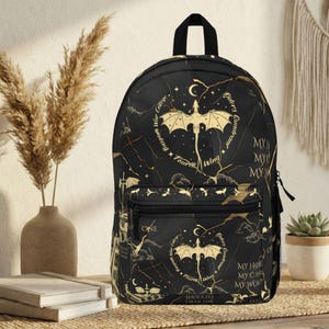 Może przedstawiać: Czarny plecak ze złotym wzorem smoka i tekstu. Plecak posiada górny uchwyt, przednią kieszeń i boczną kieszeń z siatki. Projekt zawiera smoka, księżyc, gwiazdy i tekst.