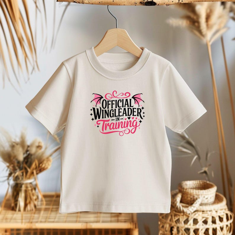 May include: A cream-colored t-shirt with the text "OFFICIAL WINGLEADER IN TRAINING" in black and pink. The design includes wing-like shapes and decorative elements. The shirt is displayed on a wooden hanger.