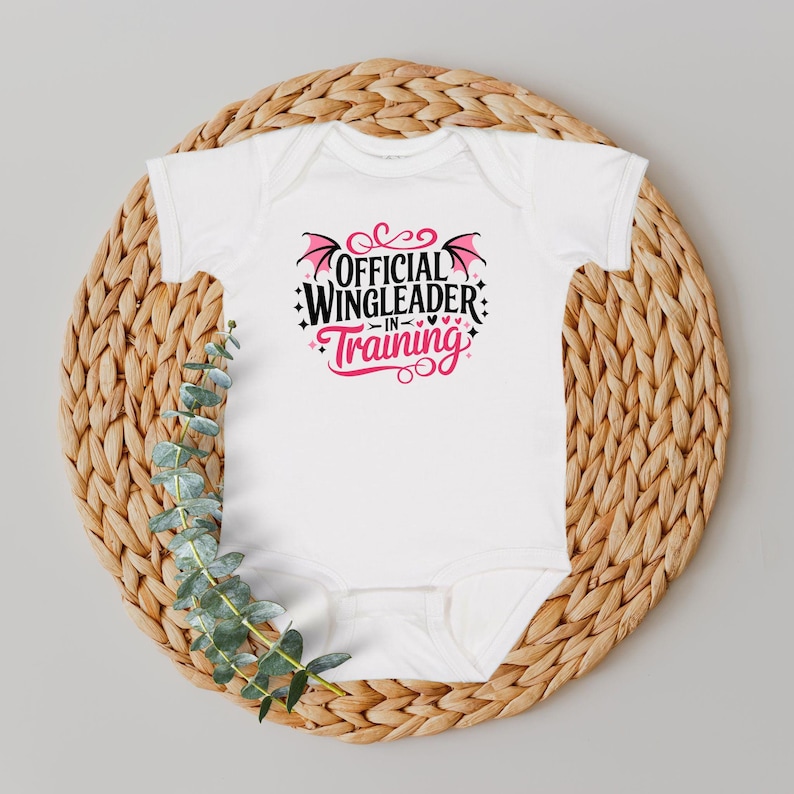 May include: White baby onesie with the text "OFFICIAL WINGLEADER IN TRAINING" in black and pink. The design includes bat wings and hearts. The onesie is displayed on a woven, circular placemat with green foliage.