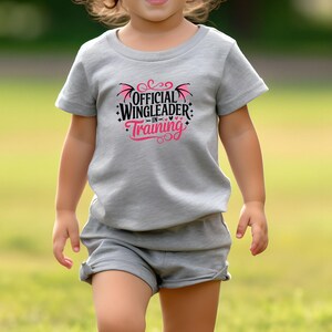 May include: A gray t-shirt and shorts set with the text "OFFICIAL WINGLEADER IN TRAINING" in black and pink. The shirt features a graphic of wings and hearts. The outfit is for children.