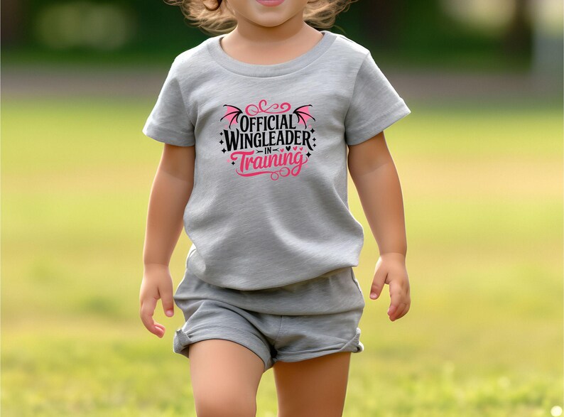 May include: A gray t-shirt and shorts set with the text "OFFICIAL WINGLEADER IN TRAINING" in black and pink. The shirt features a graphic of wings and hearts. The outfit is for children.
