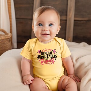 May include: A smiling baby wearing a yellow onesie with the text "OFFICIAL WINGLEADER -IN- TRAINING" in black and pink. The onesie has short sleeves and is made of a soft material. The baby is sitting on a beige blanket.