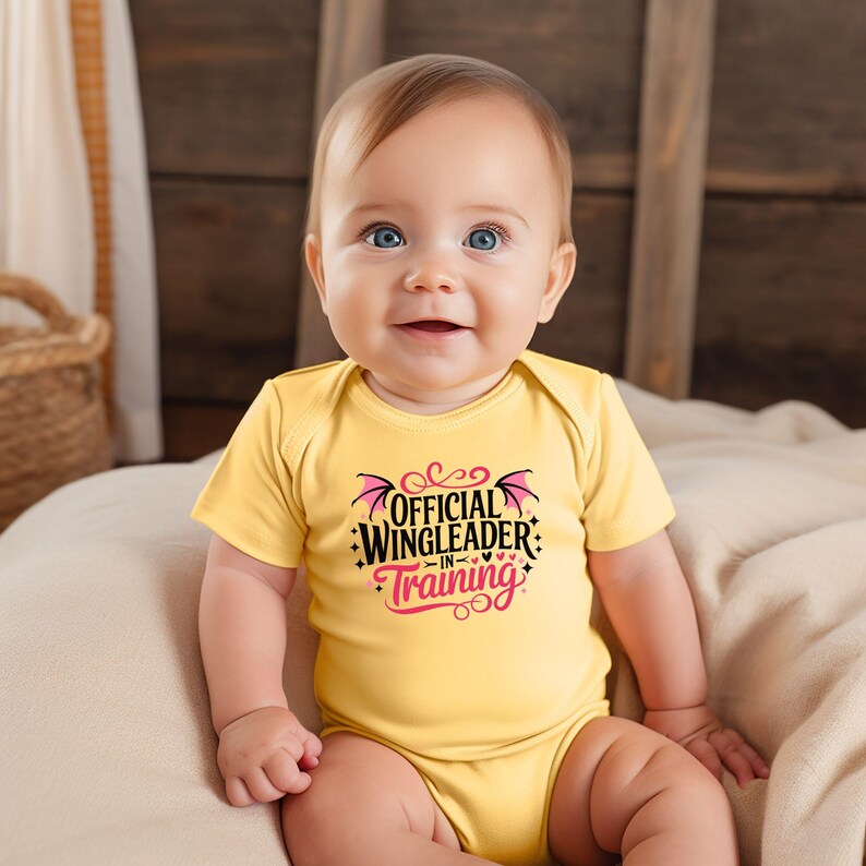 May include: A smiling baby wearing a yellow onesie with the text "OFFICIAL WINGLEADER -IN- TRAINING" in black and pink. The onesie has short sleeves and is made of a soft material. The baby is sitting on a beige blanket.
