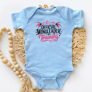 May include: A light blue baby bodysuit with the text "OFFICIAL WINGLEADER IN TRAINING" in black and pink. The design includes wing and heart graphics. The bodysuit is displayed on a woven, beige mat with a wooden bead garland.