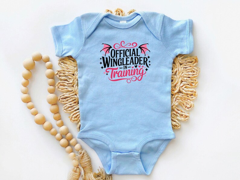May include: A light blue baby bodysuit with the text "OFFICIAL WINGLEADER IN TRAINING" in black and pink. The design includes wing and heart graphics. The bodysuit is displayed on a woven, beige mat with a wooden bead garland.