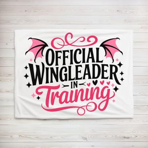 May include: White blanket with the text "OFFICIAL WINGLEADER IN TRAINING" in black and pink. The design includes pink bat wings, hearts, and stars. The blanket is displayed on a light wood background.