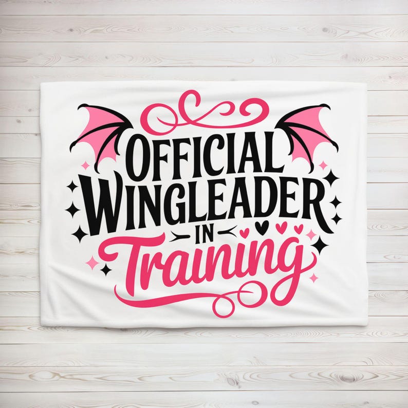 May include: White blanket with the text "OFFICIAL WINGLEADER IN TRAINING" in black and pink. The design includes pink bat wings, hearts, and stars. The blanket is displayed on a light wood background.
