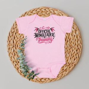 May include: A light pink baby bodysuit with the text "OFFICIAL WINGLEADER IN TRAINING" in black and red. The bodysuit is displayed on a woven wicker placemat with green foliage.
