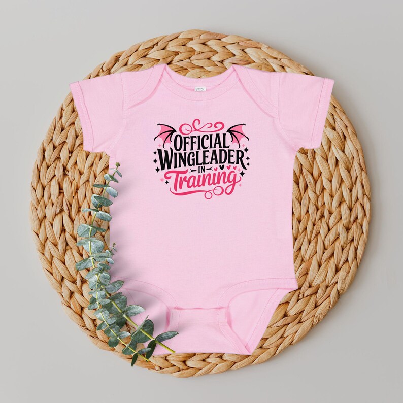 May include: A light pink baby bodysuit with the text "OFFICIAL WINGLEADER IN TRAINING" in black and red. The bodysuit is displayed on a woven wicker placemat with green foliage.