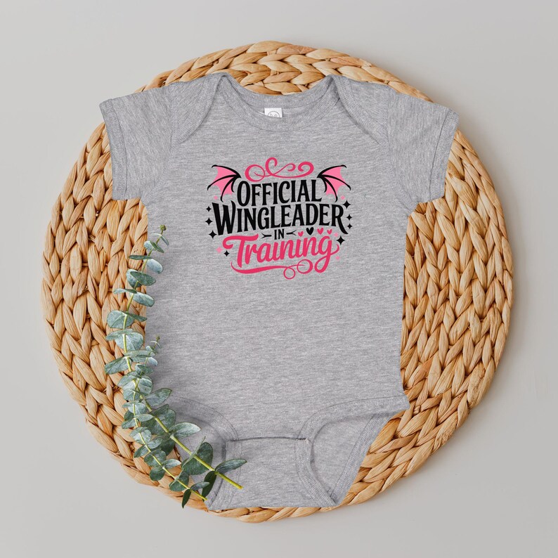 May include: A gray baby onesie with the text "OFFICIAL WINGLEADER IN TRAINING" in black and pink. The onesie is displayed on a round, woven wicker placemat with a sprig of green leaves.