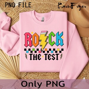Może przedstawiać: R&oacute;żowa bluza z nadrukiem graficznym, kt&oacute;ry m&oacute;wi "Rock the Test" kolorowymi literami z błyskawicą i oł&oacute;wkiem.