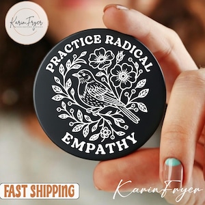 May include: Black round phone grip with the words "PRACTICE RADICAL EMPATHY" in white. A white bird and floral design is in the center. The grip is held in a hand.