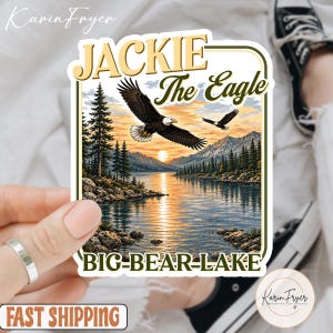 May include: A rectangular sticker featuring a landscape illustration of Big Bear Lake at sunset with two bald eagles flying above the water. The text reads JACKIE The Eagle at the top and BIG-BEAR-LAKE at the bottom in dark green lettering.