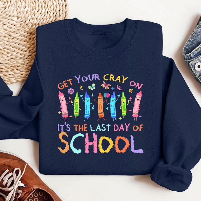 May include: Navy blue sweatshirt with a colorful crayon graphic. The text reads "Get Your Cray On" above a row of animated crayons and "It's The Last Day of School" below. A fun design for back-to-school or end-of-year celebrations.