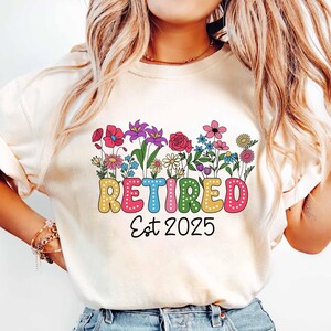 Puede incluir: Una camiseta blanca con un dise&ntilde;o floral colorido y el texto "Retired Est 2025".