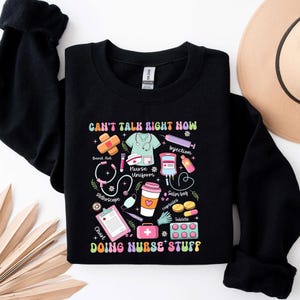 Puede incluir: Sudadera negra con un diseño gráfico colorido de suministros médicos y el texto "Can't talk right now doing nurse stuff".