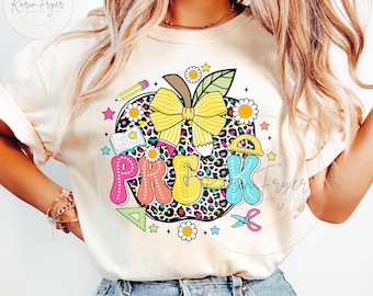 May include: A white t-shirt with a colorful graphic of an apple with the word "PRE-K" in a rainbow font. The apple is decorated with a yellow bow, a pencil, a book, a ruler, and a pair of scissors.
