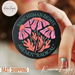 Könnte beinhalten: Schwarzer runder Handyhalter mit dem Text "BASIC HUMAN RIGHTS ARE NOT 'RADICAL'" und einem floralen Design in Pink, Koralle und Türkis. Der Halter wird in einer Hand mit einer türkis-braunen Maniküre gehalten.