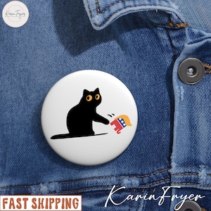 May include: A white button featuring a black cat with yellow eyes swatting at a red elephant with a blonde toupee. The button is pinned to a blue denim jacket. The words "FAST SHIPPING" are printed on the bottom left.