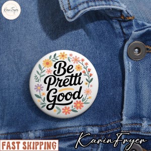 May include: A white button pin with the words "Be Pretti Good" in black script, surrounded by floral illustrations in yellow, pink, and orange. The pin is set against a blue denim jacket.