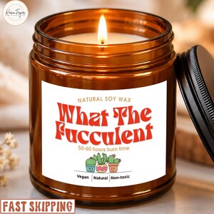 May include: A lit candle in an amber glass jar with a black lid. The white label features the red text What The Fucculent, Natural Soy Wax, 50-60 hours burn time, and an illustration of three potted cacti with Vegan, Natural, Non-toxic text.