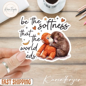 Puede incluir: Una pegatina con el texto "be the softness that the world needs", que presenta un orangután abrazando a un mono. La pegatina tiene un fondo blanco con ilustraciones en naranja, marrón y negro. La pegatina está sujeta por una mano con un anillo plateado.