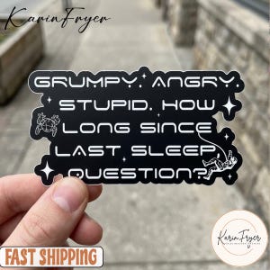 May include: A black sticker with white text that reads "GRUMPY. ANGRY. STUPID. HOW LONG SINCE LAST SLEEP QUESTION?" The sticker has a frog and astronaut graphic. The sticker is held by a hand.