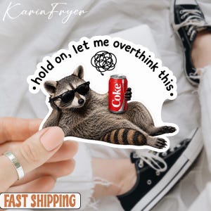 May include: A die-cut sticker featuring a brown and grey raccoon wearing black sunglasses while lying down and holding a red soda can. Above the raccoon is a black scribble icon and the text, hold on, let me overthink this, in black font.