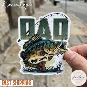 May include: A sticker featuring a detailed illustration of a largemouth bass leaping from the water, with the word "DAD" in a green, textured font above. A fishing rod is arched over the word. The sticker has a white border.