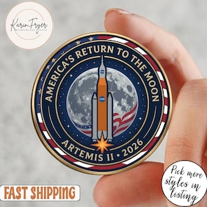 May include: A round, navy blue coin with gold trim, featuring a rocket ship design and the text "AMERICA'S RETURN TO THE MOON ARTEMIS II • 2026." The coin has a red, white, and blue border, and a moon graphic in the center.