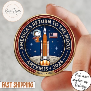 May include: A round, commemorative coin featuring the text "America's Return to the Moon" and "Artemis + 2026". The coin has a blue background with a rocket, Snoopy, and the American flag. The coin is held in a hand.